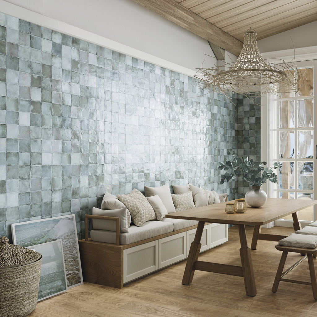 Modern rustic dining room featuring light color wood floors, wooden table, beige cushioned bench, and tiled accent wall in blue-green 4x4 ceramic tiles with  ample gray variation and fading.